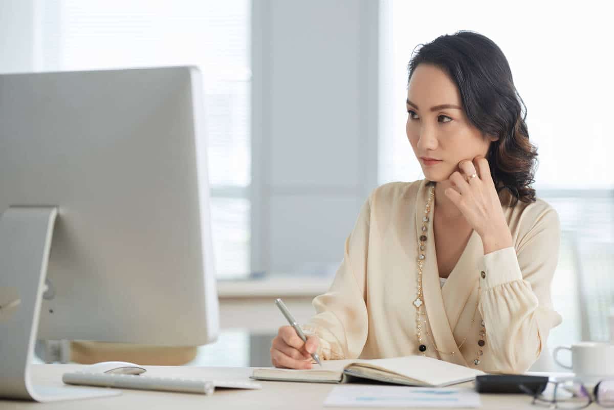 Pensive business woman reading e-mails on computer and taking notes
